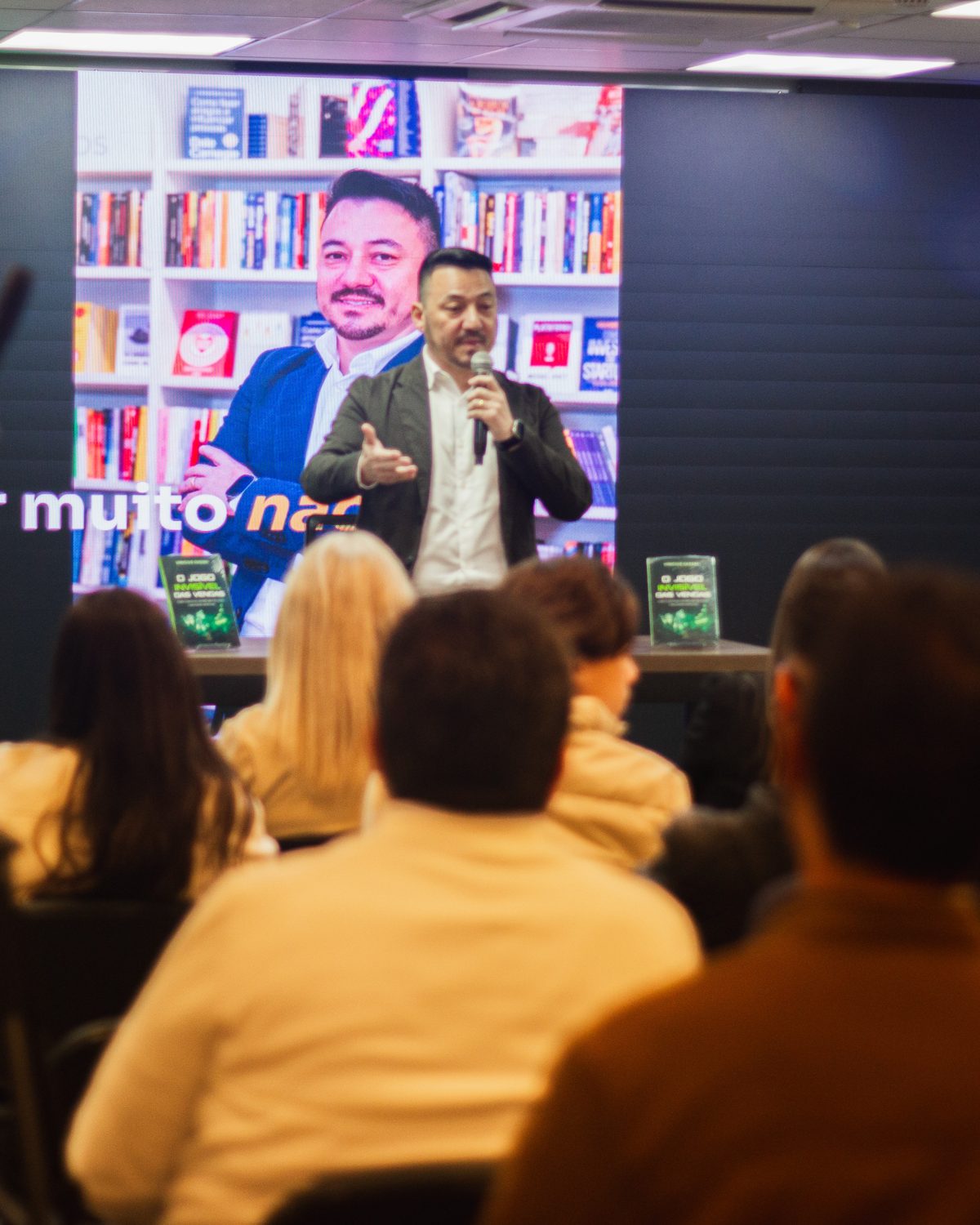 Público assiste à palestra de lançamento do livro, com Vinicius Sasaki de microfone na mão diante do telão