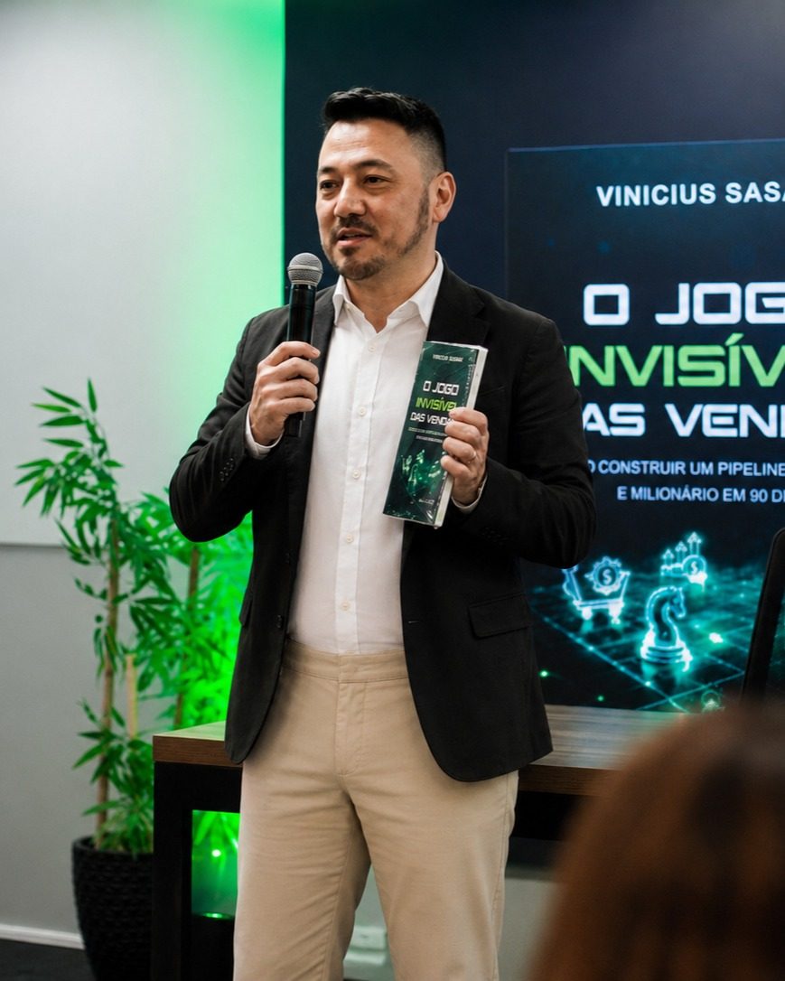 Vinicius Sasaki fala ao microfone segurando o livro O Jogo Invisível das Vendas, com a capa projetada ao fundo