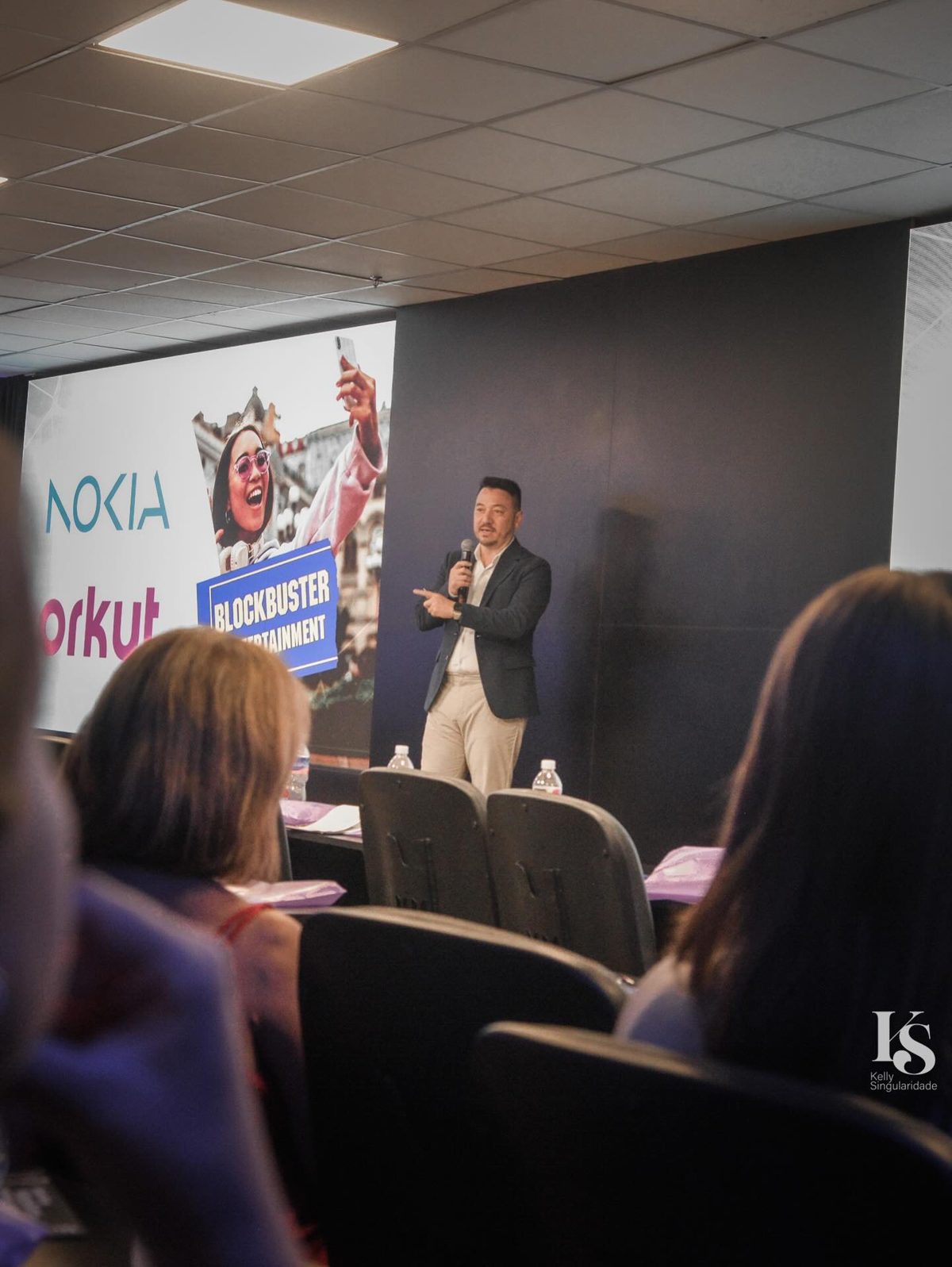 Plateia vista de trás assiste a Vinicius Sasaki; no telão, as marcas Nokia, Orkut e Blockbuster