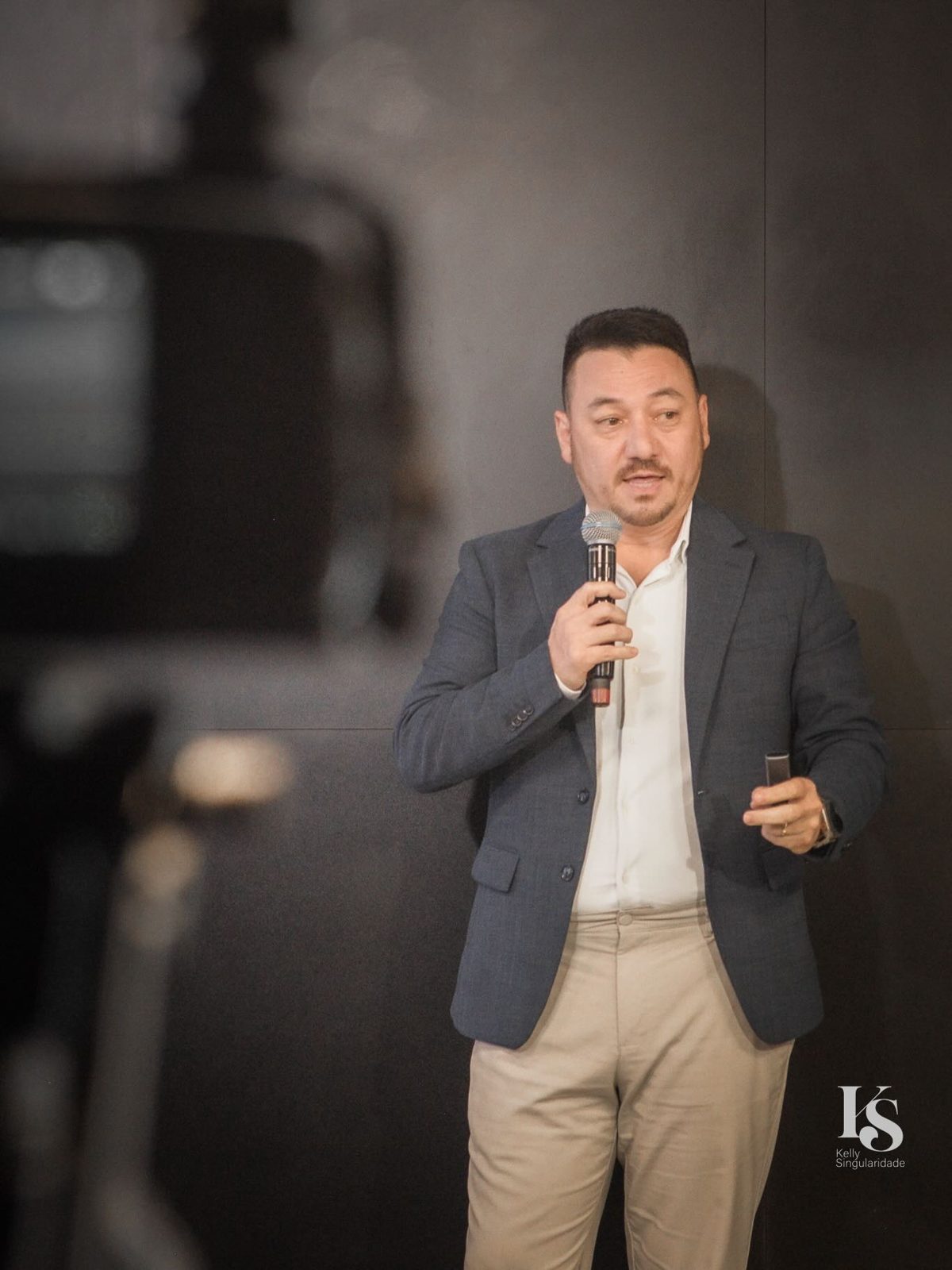 Vinicius Sasaki fala ao microfone, de pé, com o controle dos slides na outra mão